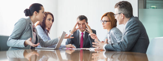 Difficult meeting with multiple people taking at one and a man in the center holding his head.