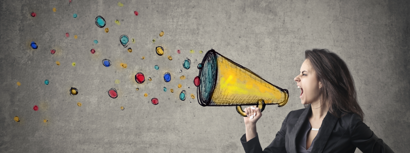Women shouting into a cartoon megaphone that is painted on a grey wall. The woman is wearing a suit.