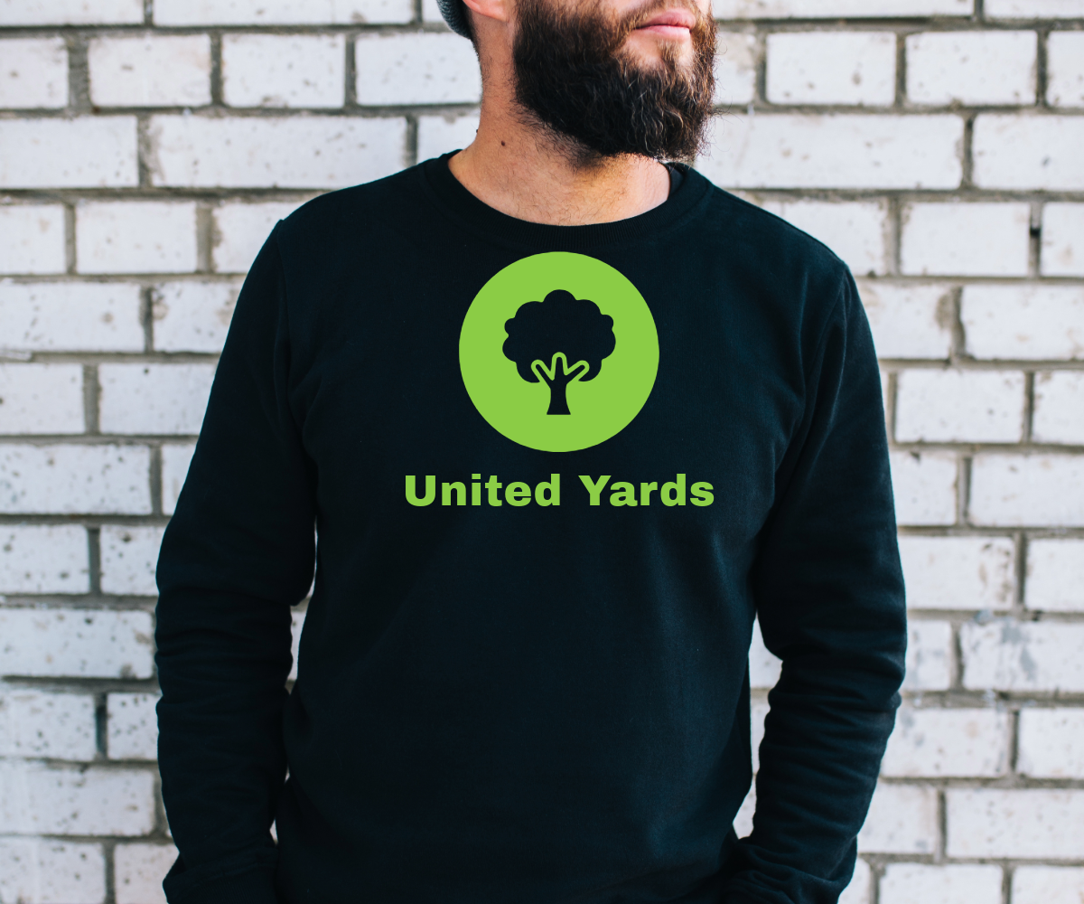 A printed shirt with united yards in green printed on it with a green circle and an outline of a tree inside of it. A man is wearing the shirt in front of a white brick wall.