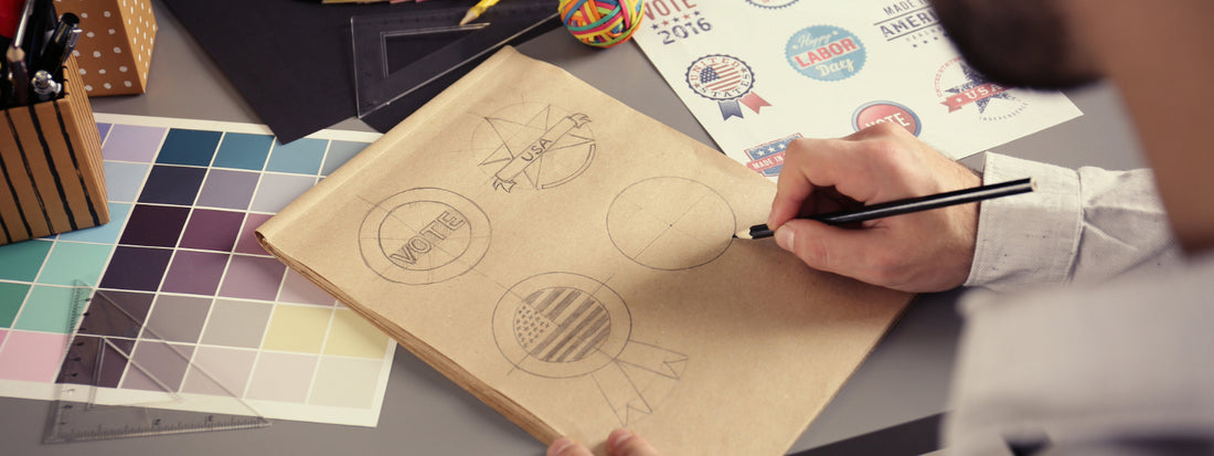 A man draws a few logos by hand on paper while looking at printed examples.