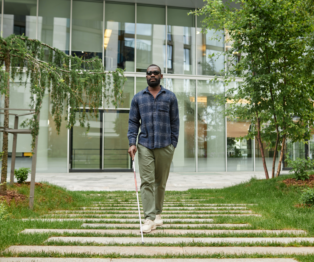 A blind man walks on a outdoor walkway glossed over with light grass. He is using a white cane to find his way, wearing a blue flannel shirt and green pants. He is walking in the center of a corporate park.