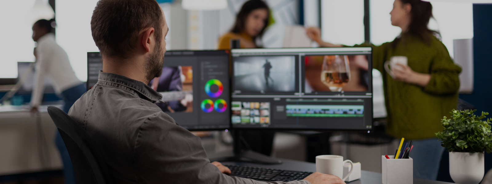 A man sits at a desk with adobe premier and designs on his monitors. Two women in the background are talking in front of him.